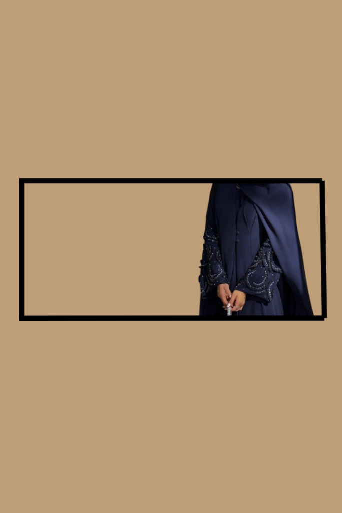 A woman wearing a navy blue abaya with intricate beadwork on the sleeves, standing against a peach-colored background with a black rectangular border.