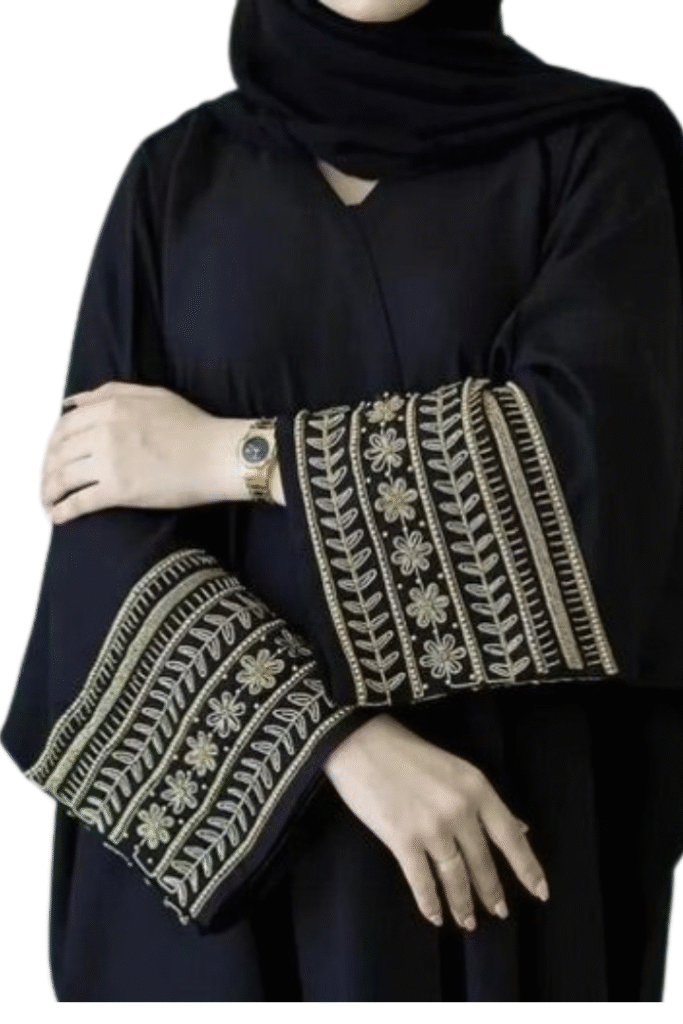 A woman wearing a black abaya with intricate gold and white embroidery on the wide sleeves, paired with a black headscarf. She is wearing a gold watch and a ring