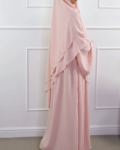 Woman wearing a light pink layered abaya and matching khimar, standing sideways against a white wall.