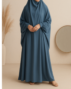 A woman wearing a slate blue jilbab and matching hijab stands gracefully in a minimalist studio with pampas grass and a round mirror in the background.