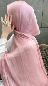 Close-up of a person wearing a pale pink, slightly shiny or crushed-effect satin hijab, elegantly draped around the neck and shoulders. The person's hand, adorned with large, textured silver rings, is visible near the chin.