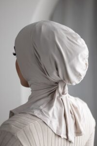 Rear view of a person wearing a close-fitting, beige/nude hijab cap/underscarf made from a soft jersey-like material, tied neatly at the nape of the neck.