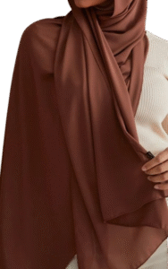 wearing a smooth, matte dark brown (chocolate) hijab draped over a ribbed, cream-colored top. A hand subtly holds the edge of the fabric.