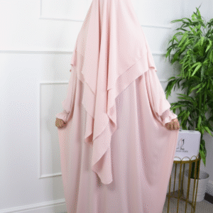 Woman wearing a light pink layered abaya and matching khimar, standing sideways against a white wall.