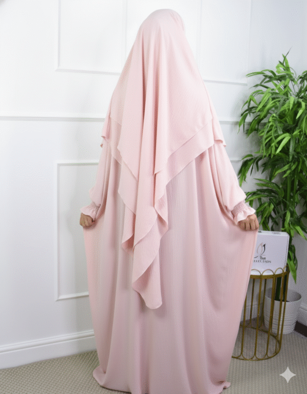 Woman wearing a light pink layered abaya and matching khimar, standing sideways against a white wall.