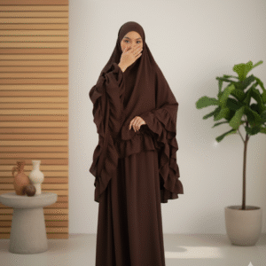 Aesthetically styled photo of a model in a voluminous brown jilbab, sitting composedly on a sleek white chair in a studio. The backdrop features abstract pastel shapes in warm, neutral colors.