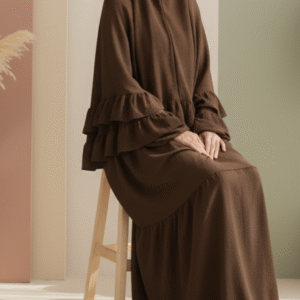 Aesthetically styled photo of a model in a voluminous brown jilbab, sitting composedly on a sleek white chair in a studio. The backdrop features abstract pastel shapes in warm, neutral colors.