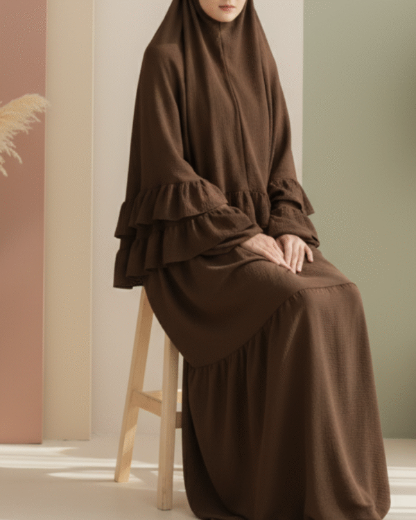 Aesthetically styled photo of a model in a voluminous brown jilbab, sitting composedly on a sleek white chair in a studio. The backdrop features abstract pastel shapes in warm, neutral colors.