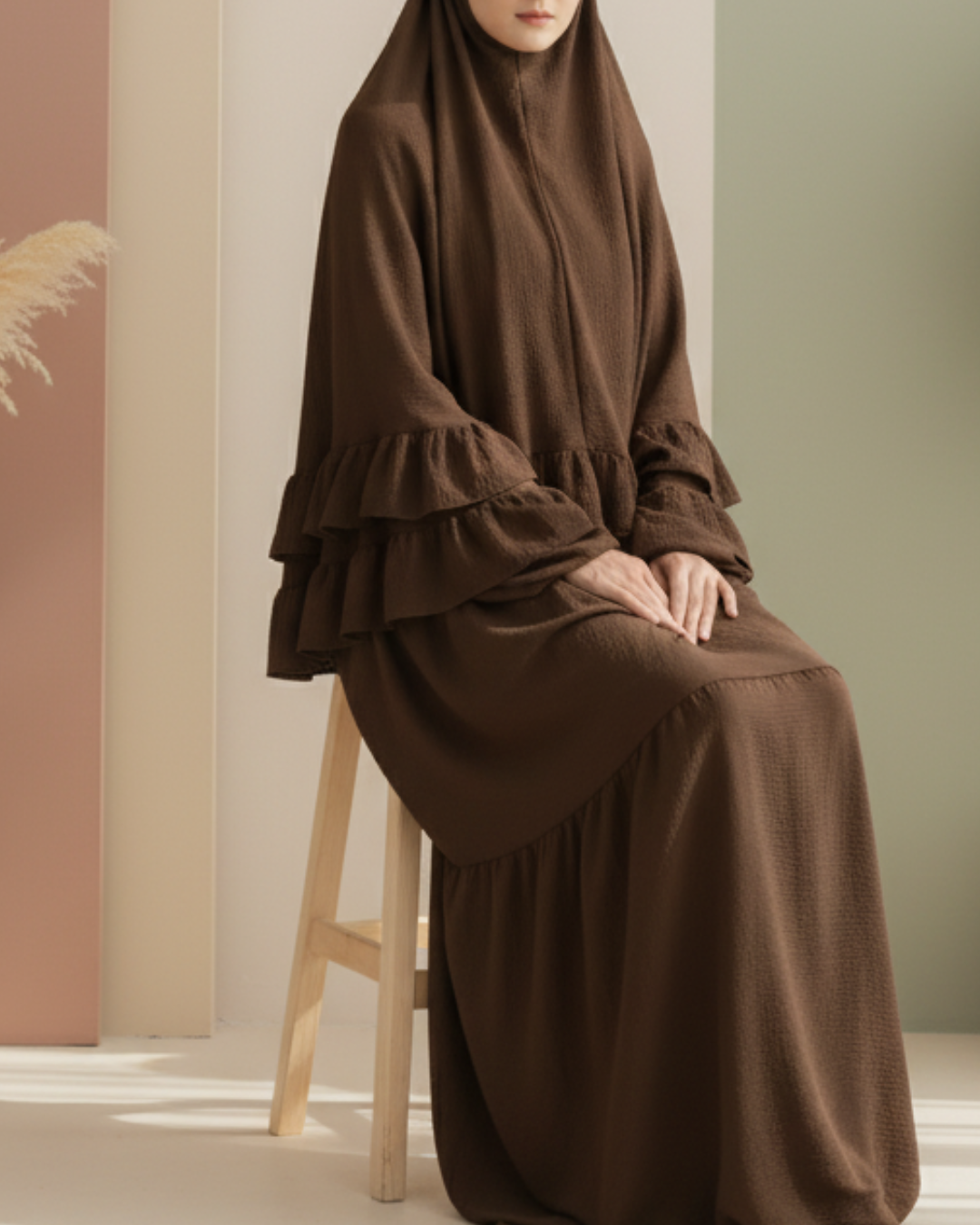Aesthetically styled photo of a model in a voluminous brown jilbab, sitting composedly on a sleek white chair in a studio. The backdrop features abstract pastel shapes in warm, neutral colors.