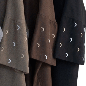 wearing a brown, flowing abaya with white embroidered crescent moon designs on the sleeves. The background is a simple, warm beige, highlighting the garment