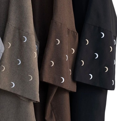 wearing a brown, flowing abaya with white embroidered crescent moon designs on the sleeves. The background is a simple, warm beige, highlighting the garment