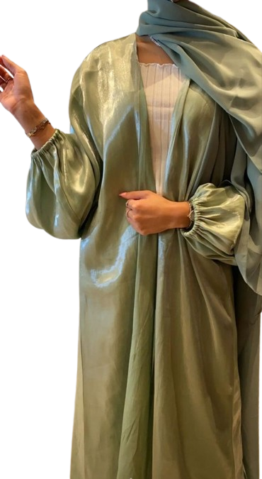 A woman in a flowing, shimmering sage green abaya with voluminous balloon sleeves, worn open over a white ribbed top and paired with a matching green hijab.