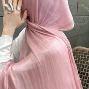 Close-up of a person wearing a pale pink, slightly shiny or crushed-effect satin hijab, elegantly draped around the neck and shoulders. The person's hand, adorned with large, textured silver rings, is visible near the chin.
