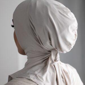 Rear view of a person wearing a close-fitting, beige/nude hijab cap/underscarf made from a soft jersey-like material, tied neatly at the nape of the neck.