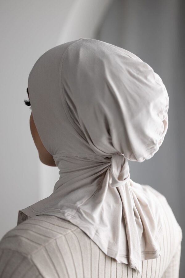 Rear view of a person wearing a close-fitting, beige/nude hijab cap/underscarf made from a soft jersey-like material, tied neatly at the nape of the neck.