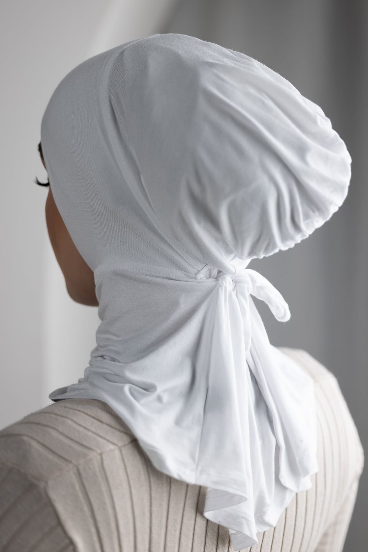Rear view of a person wearing a close-fitting, beige/nude hijab cap/underscarf made from a soft jersey-like material, tied neatly at the nape of the neck.