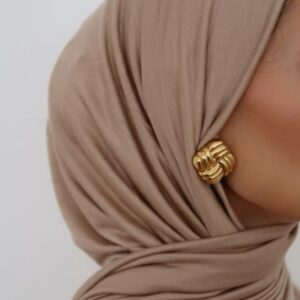 Close-up profile view of a person wearing a soft, draped, sand-colored jersey knit hijab, secured with a large, textured gold knot earring visible on the side.
