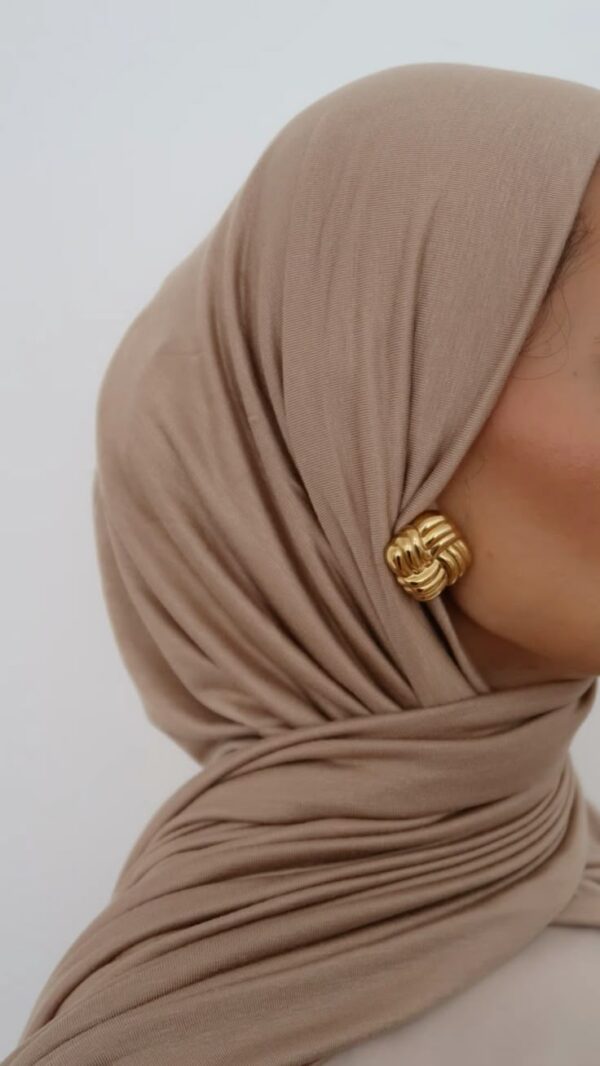 Close-up profile view of a person wearing a soft, draped, sand-colored jersey knit hijab, secured with a large, textured gold knot earring visible on the side.